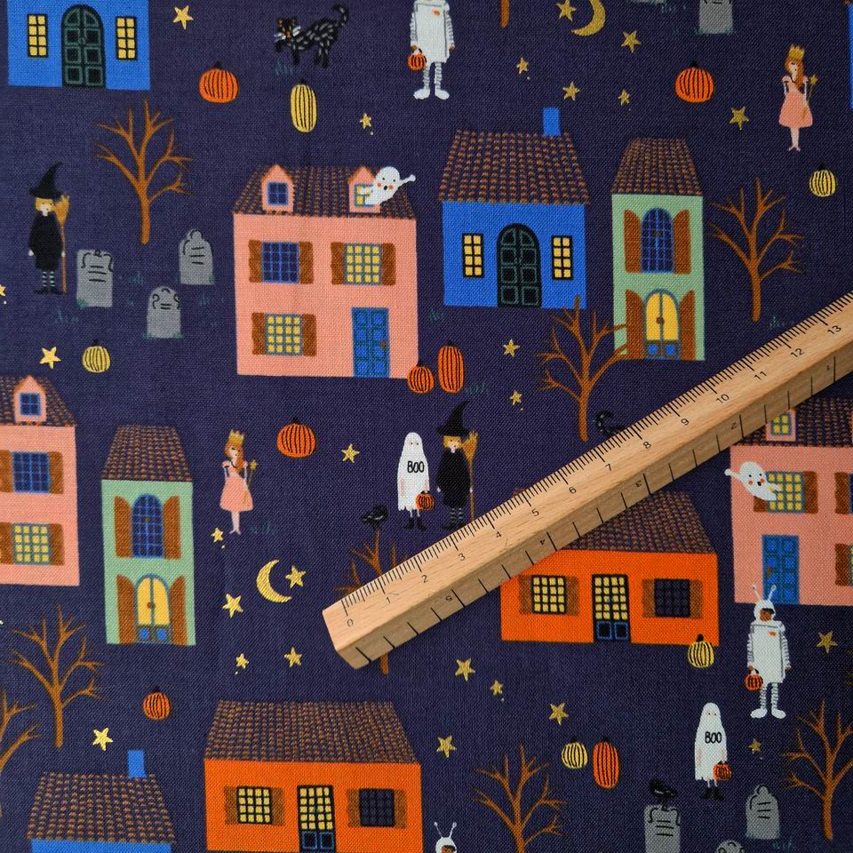 Tissu coton Rifle paper Co imprimé sur fond bleu nuit des maisonnettes éclairées bleues, vertes, et rose, des citrouilles oranges, des lunes et des étoiles comme un village la nuit d'halloween
